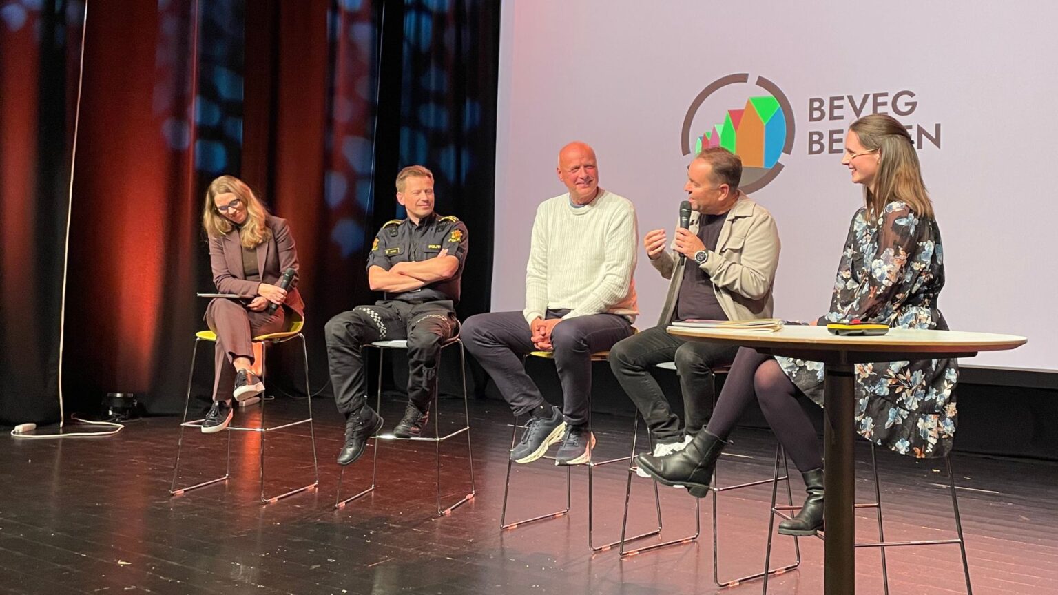 Fem mennesker i panelsamtale på scene, med logo til lansering av Beveg Bergen i bakgrunn.